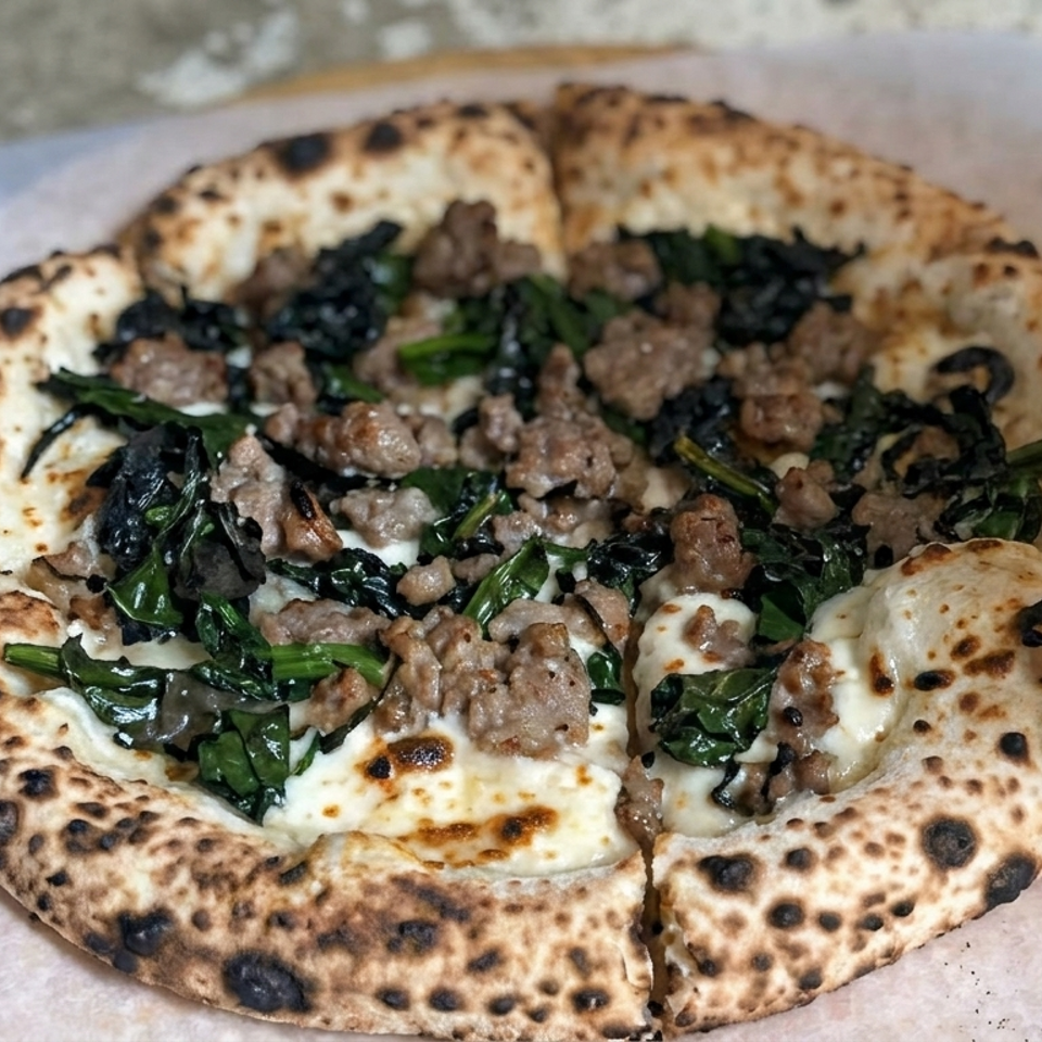 Neapolitan-style pizza with charred crust, sausage, leafy greens, mushrooms, and melted mozzarella on parchment paper.
